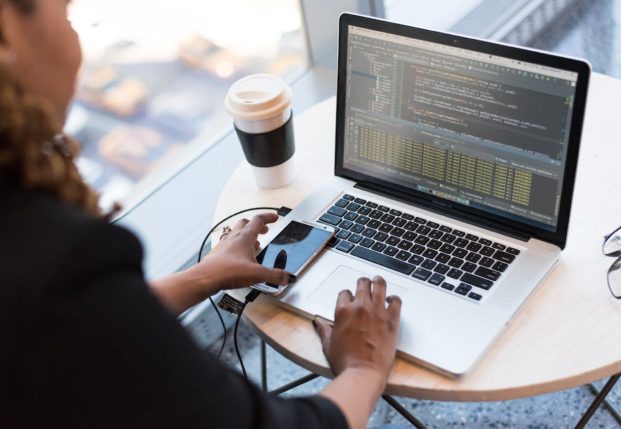 A woman sits at her laptop looking at code on her screen. Her left hand is reaching for her mobile telephone. A woman sits at her laptop looking at code on her screen. Her left hand is reaching for her mobile telephone.