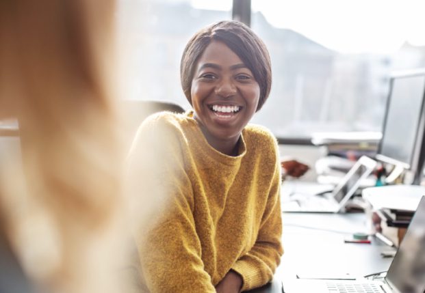 Woman smiling at another co-worker sitting in front of desk Woman smiling at another co-worker sitting in front of desk